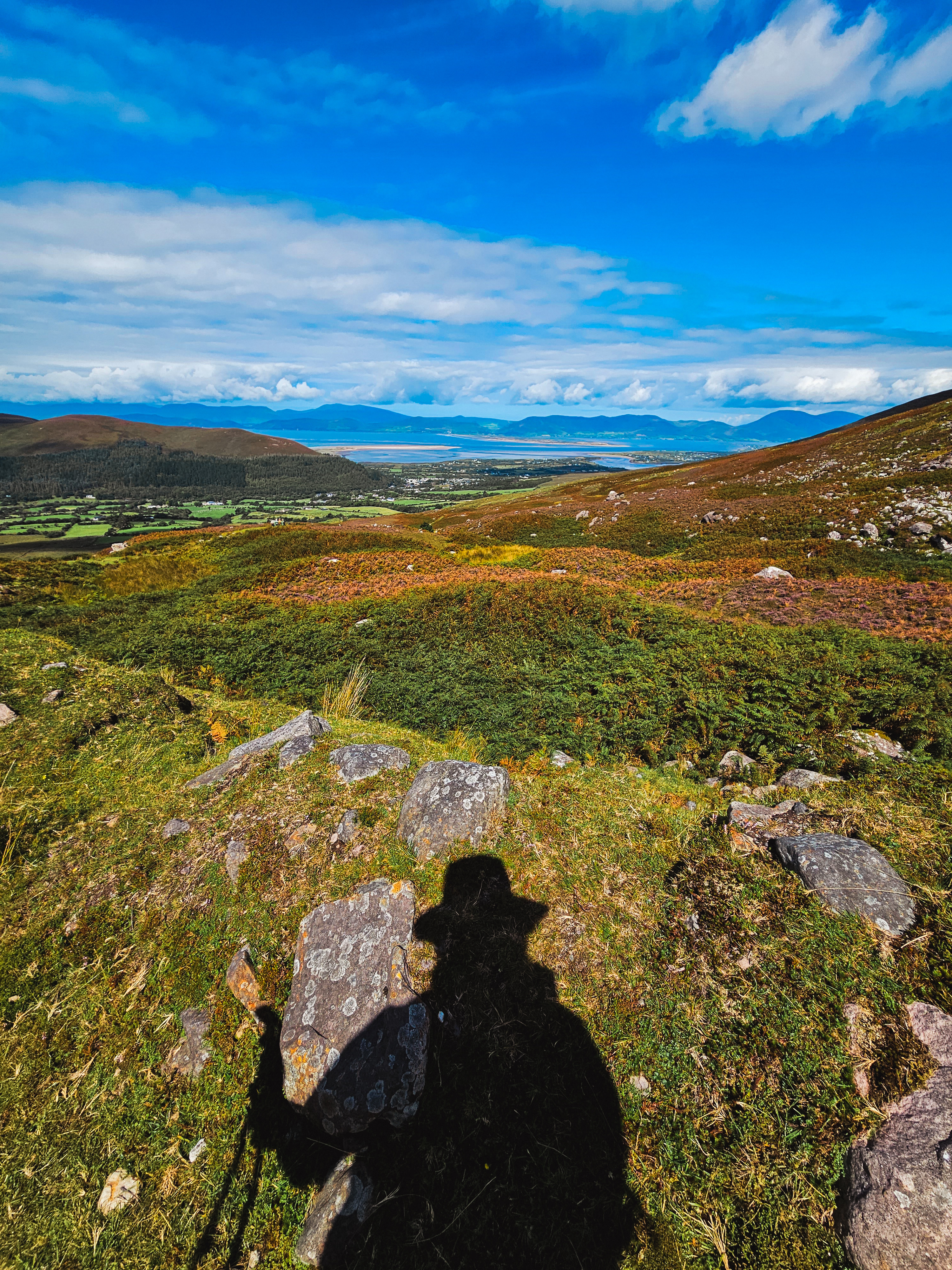 Bir dağ manzarasıyla birlikte, ön planda bir yürüyüşçünün gölgesi. Yeşil araziler ve uzak dağlar görünmekte.