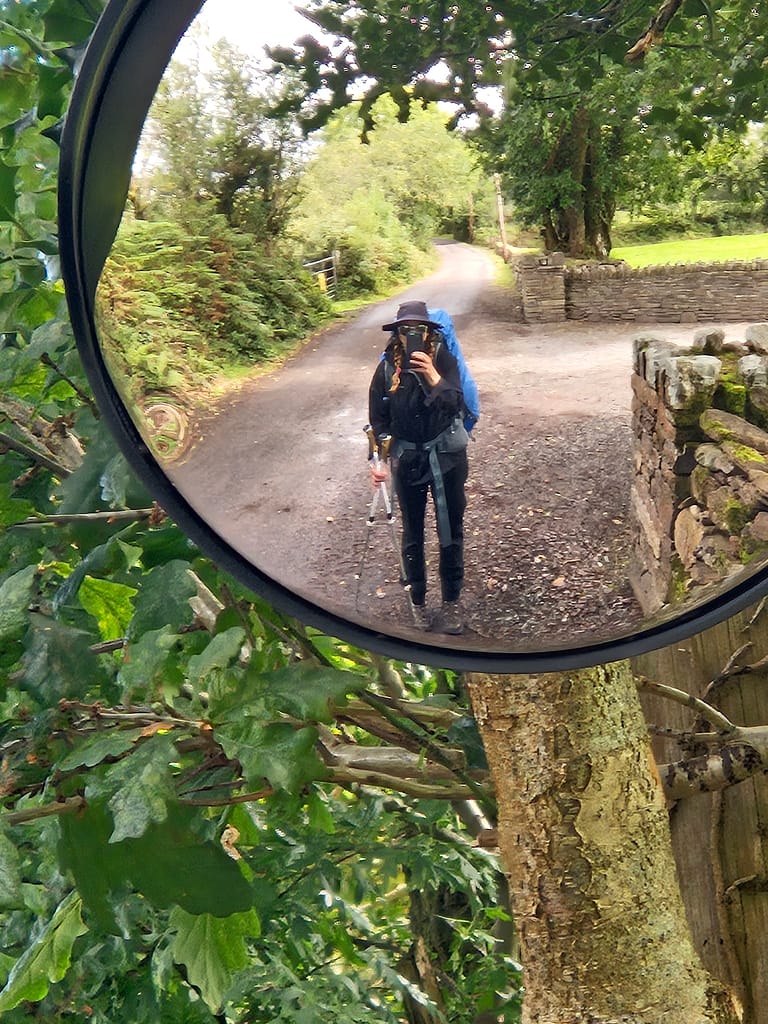 Yolda yürüyüşe çıkan bir kişinin ayna yansıması, ormanlık bir alanda yürüyüş ekipmanlarıyla birlikte görünüyor.