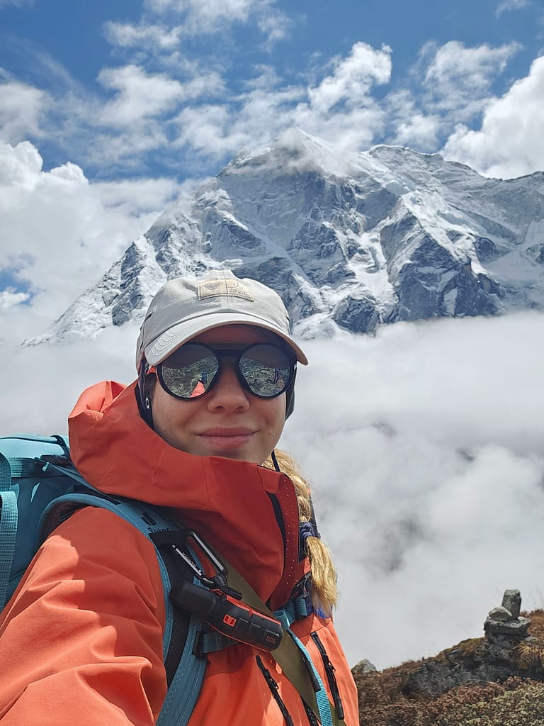 Bir dağ manzarası önünde, turuncu bir mont giymiş ve gözlük takmış bir kadın kendini çekiyor. Arka planda bulutların arasında karla kaplı zirveler görünmekte.