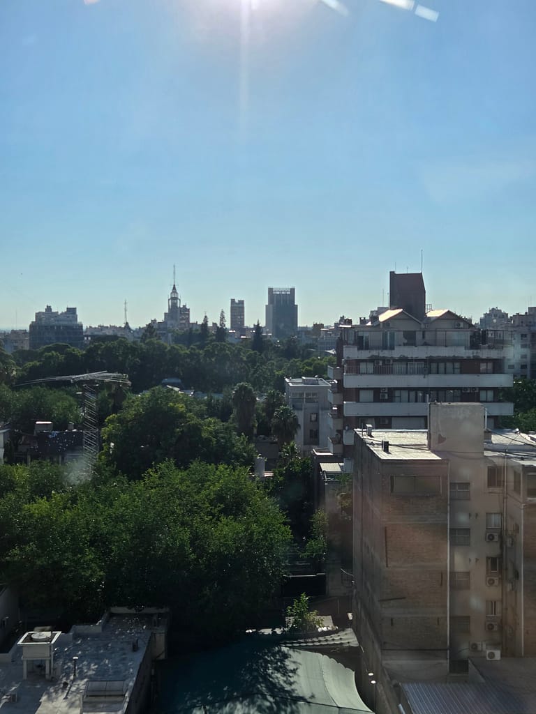 Şehir silueti üzerinde güneşin parladığı bir manzara, yeşil ağaçlar ve binalar görünmekte.