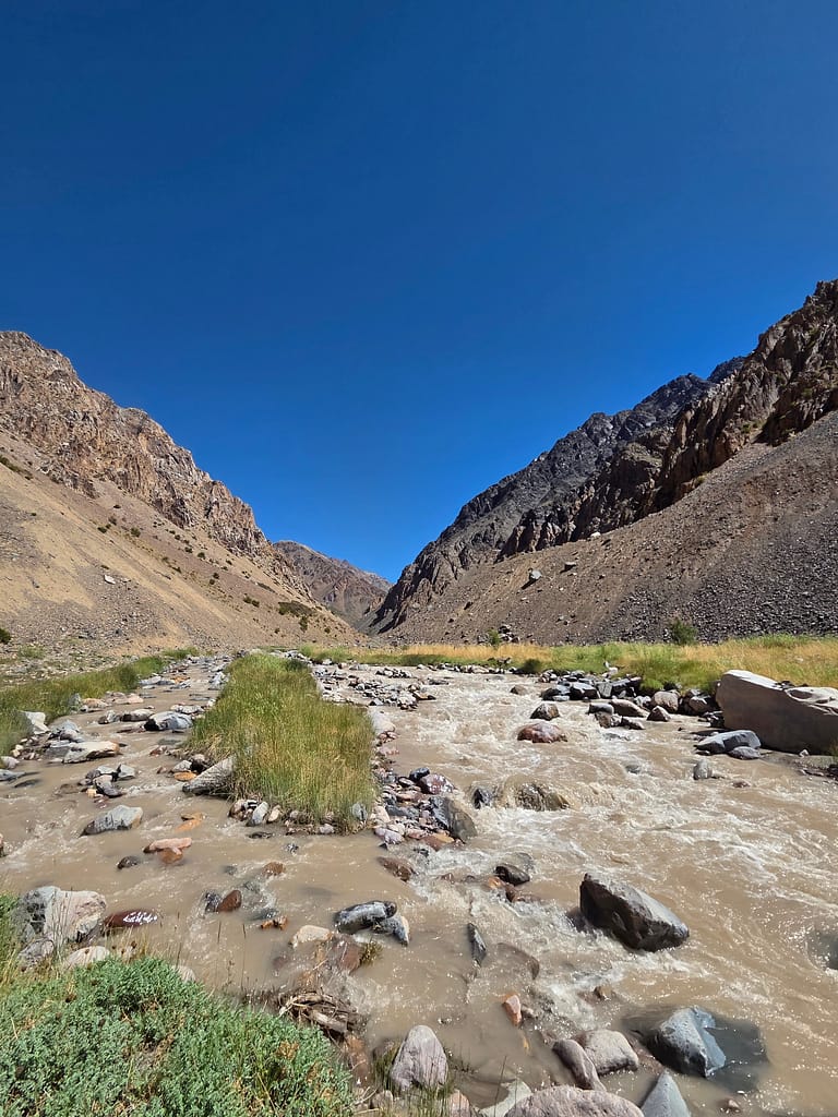 Dağlık bir vadi içerisinde akan çamurlu bir nehir, etrafında taşlar ve yeşil bitkilerle çevrili.
