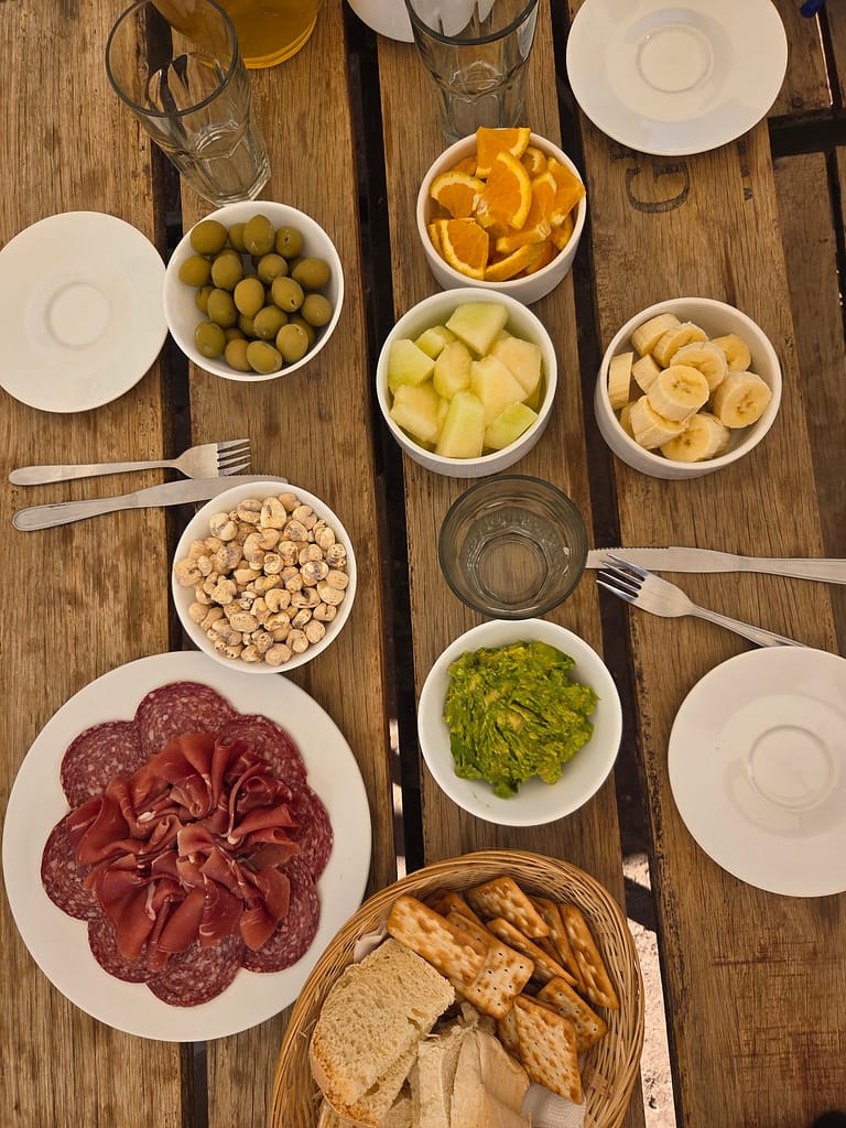 Ahşap masada çeşitli atıştırmalıklar; zeytin, meyveler, şarküteri ürünleri, kuruyemiş ve ekmek yer almakta.