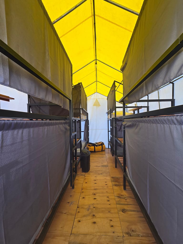 İç mekanında üst üste yatakların bulunduğu bir kamp alanı, sarı tente ile örtülü ve ahşap zeminli.