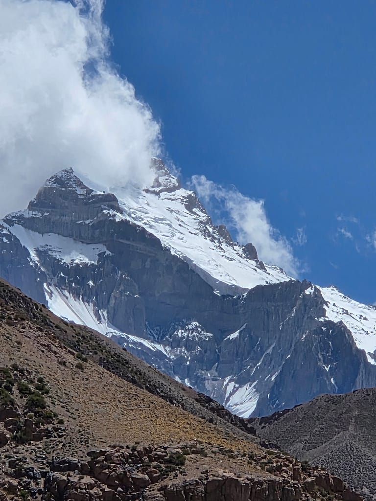 Bembeyaz karlarla kaplı yüksek dağ, mavi gökyüzü ve bulutlar arasında görünüyor.