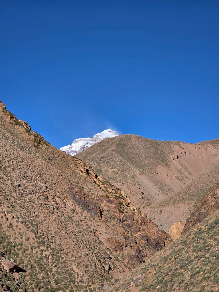 Buzul kaplı bir dağın tepe kısmının görünümü, çevresinde sarp yamaçlar ve açık mavi bir gökyüzü ile.