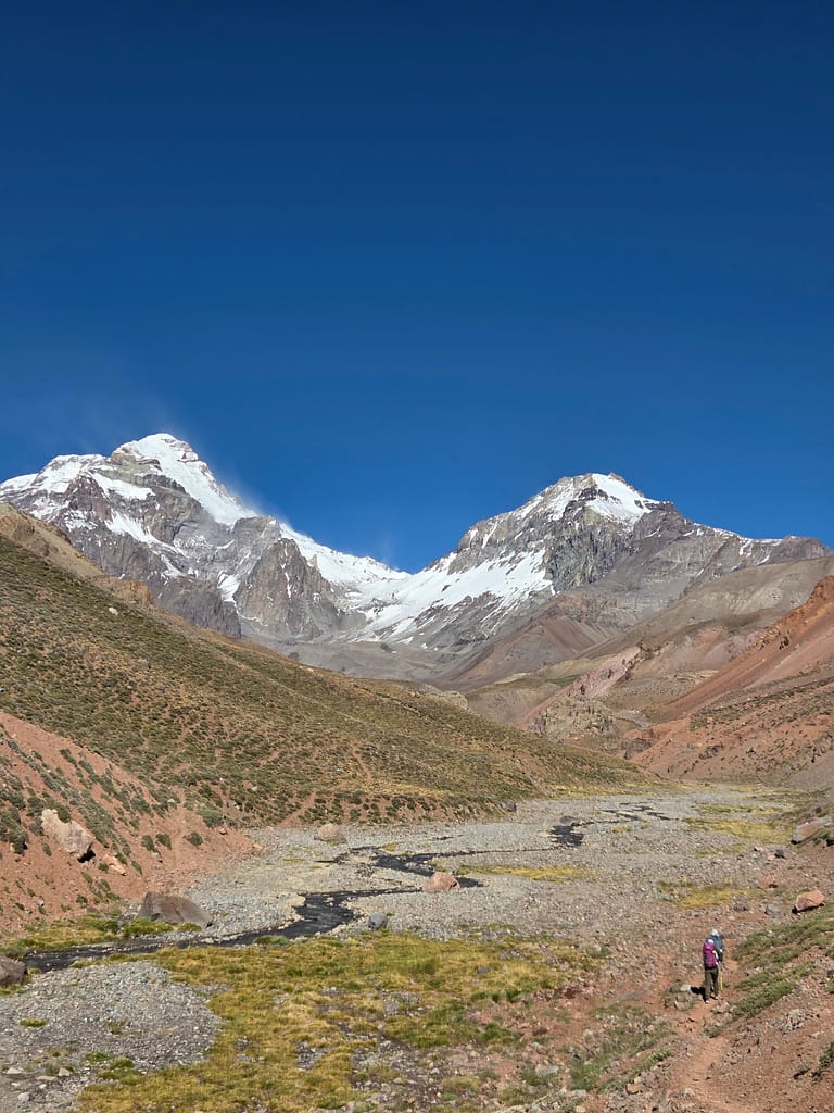 Buzullarla kaplı dağların ve yeşil alanların bulunduğu bir vadiye yürüyen bir kişinin görüntüsü.