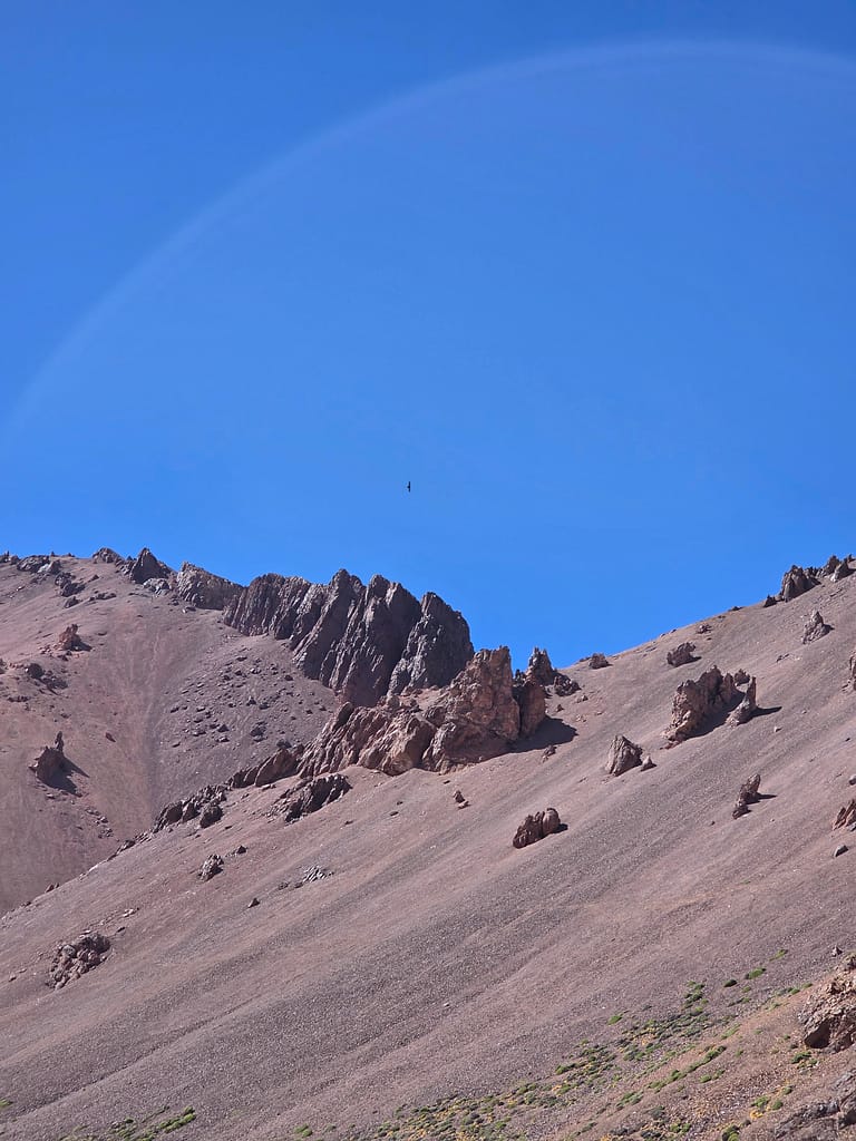 Mavi gökyüzü arka planında yer alan dik kayalıklara sahip dağ manzarası; birkaç yeşillik bitki görülüyor.