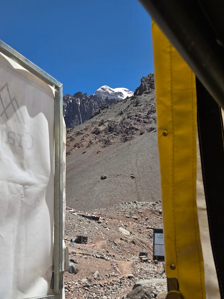 Bir dağın tepesi ve mavi gökyüzü görünümüne sahip bir manzara. Solda bir yapı yer alıyor.