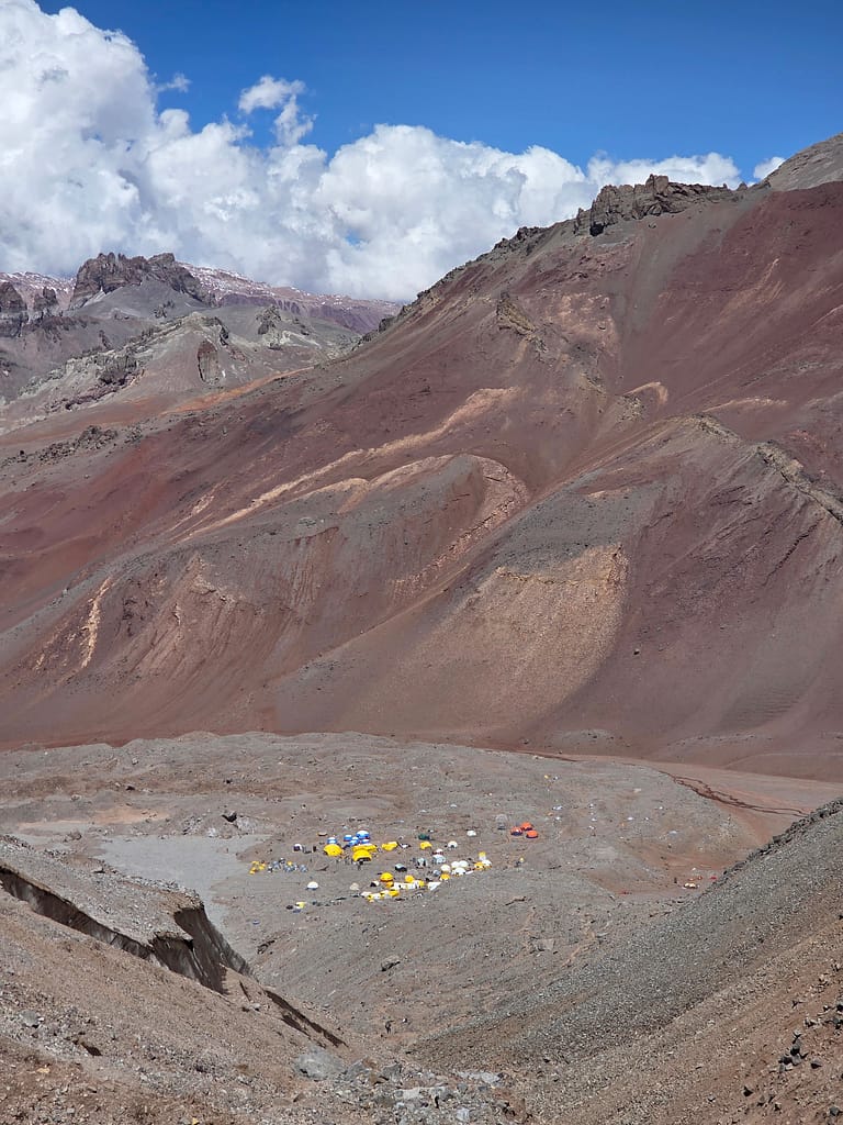 Yüksek dağlık alanda yer alan kamp alanı, sarı ve mavi çadırlarla dolu. Arka planda, yüksek dağlar ve mavi gökyüzü bulutlarla kaplı.