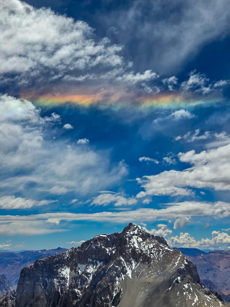 Dağ zirvesinin üzerinde oluşan bir gökkuşağı ile mavi gökyüzü ve bulutlar.