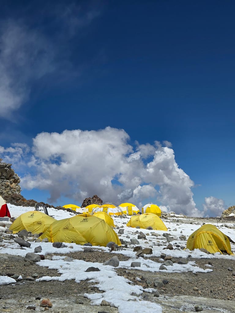 Karla kaplı dağlık alanda yer alan sarı çadırlar, mavi gökyüzü ve beyaz bulutlarla çevrili.