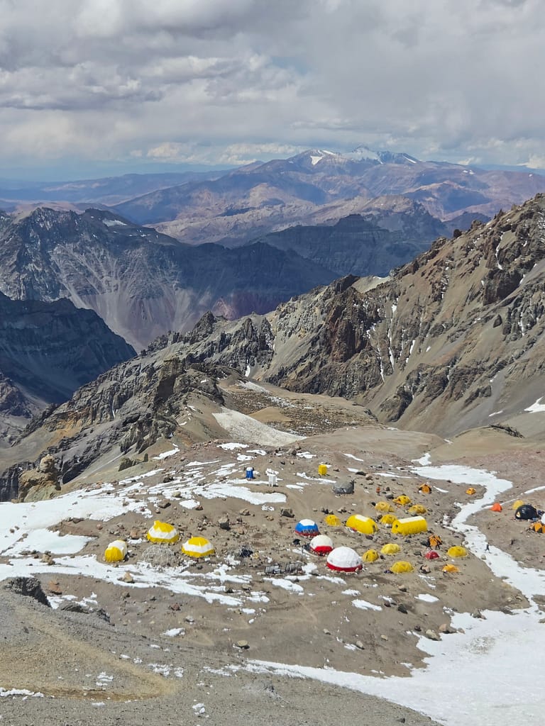 Yüksek dağ zirvesinde kurulu renkli çadırlar ve karla kaplı arazinin manzarası.