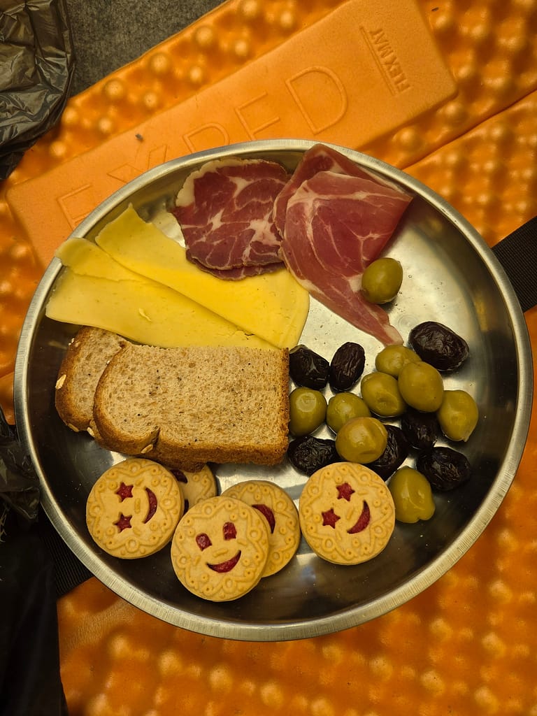 Kahvaltı tabağında iki çeşit peynir, dilimlenmiş jambon, zeytinler, kuru üzüm ve gülümseyen şekilli kurabiyeler bulunan bir metal tabak.