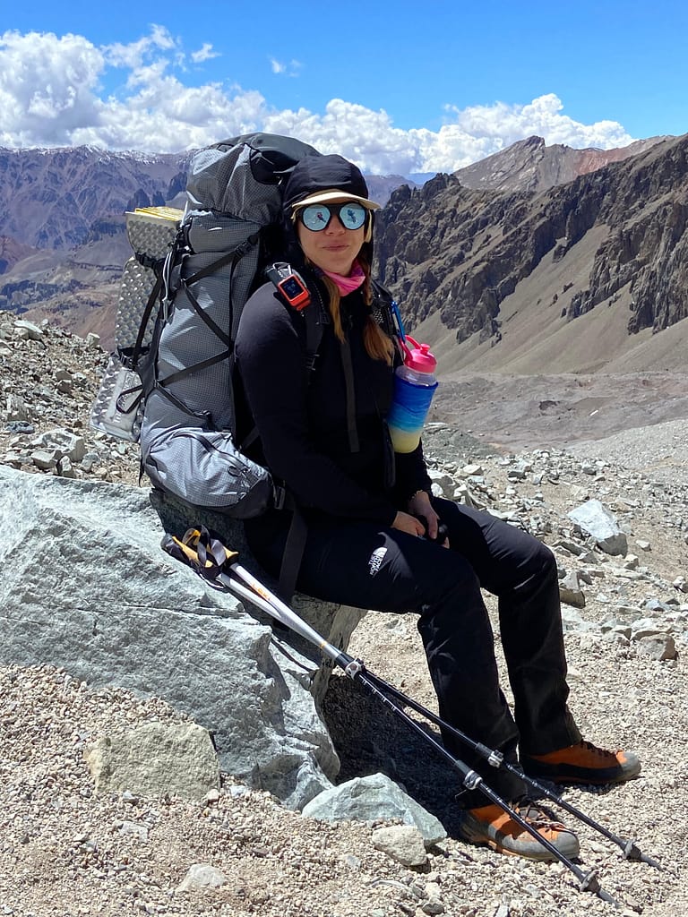 Dağlık bir alanda oturan, sırt çantası taşıyan ve güneş gözlüğü takan bir kadın.