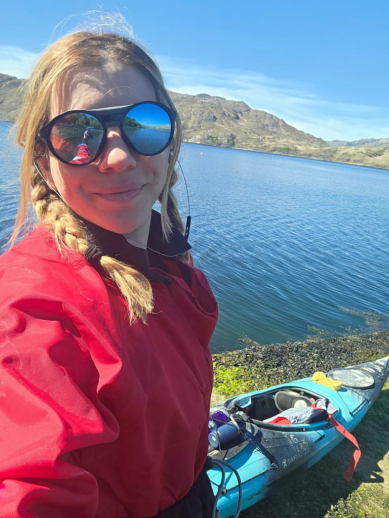 Göl kenarında bir kayak ile selfie çeken kadın, kırmızı bir giysi ve büyük gözlükler giyiyor.
