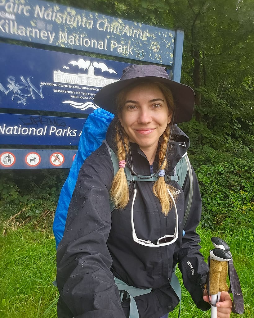 A hiker smiles in front of a sign for Killarney National Park, carrying a large pack and a walking stick.