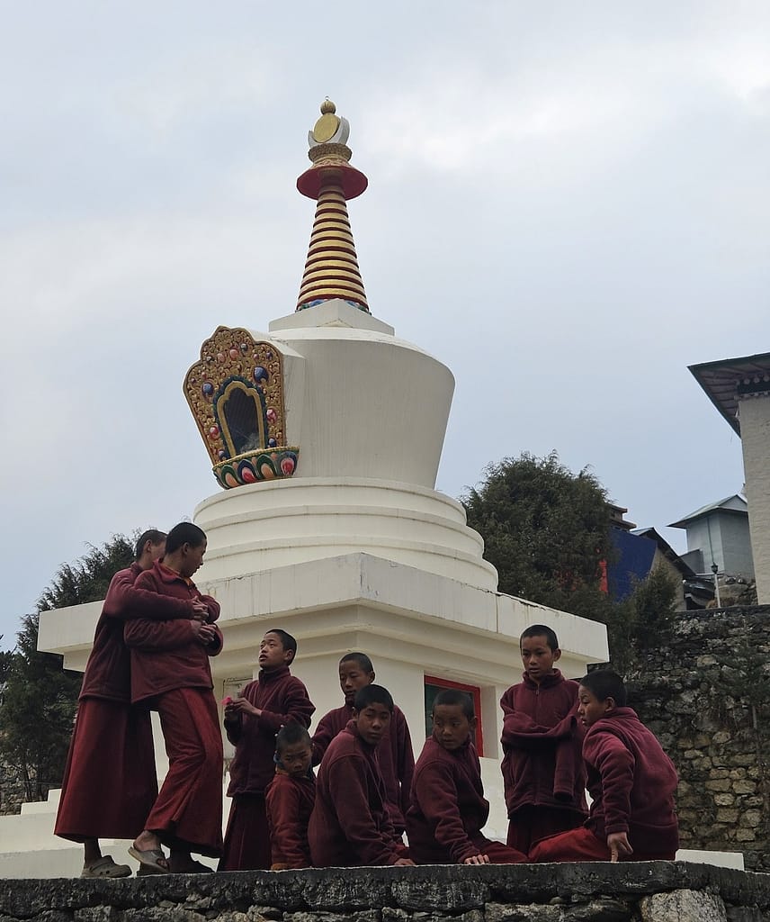 Bir grup genç keşiş, bir stupa önünde oturmuş ve sohbet ediyor. Arka planda açık gökyüzü ve çevre mimarisi yer alıyor.